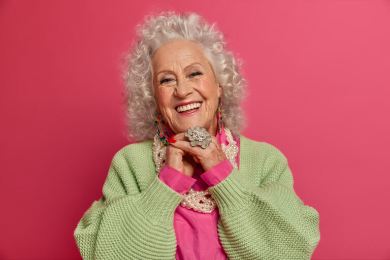 Portrait of happy grey haired wrinkled eldrely woman has well cared face, wears makeup, fashionable clothes and accessories, smiles toothily, isolated on pink background, always looks young.
